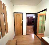 A two-room apartment with a wood-patterned floor, a coat rack wall, and open doors to a room.