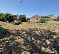 Building plot near a family house in Mužla with a grassy area, trees, and neighboring buildings.