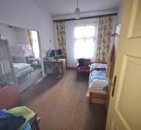 A room in a family house with a carpet, a bed, a computer, and a window with floral curtains.