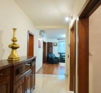A hallway in a family house with tiles and wooden furniture leads to a living room with a wood-patterned floor.