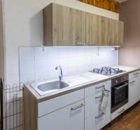 A kitchen with wood-patterned flooring, white tiles, and a gas stove in a 2-room apartment.