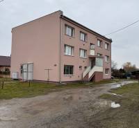 Apartment building in Ludanice with four windows and an entrance staircase on a wet road.
