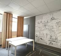 The office has white desks, a drawing on the wall, and vertical window blinds.