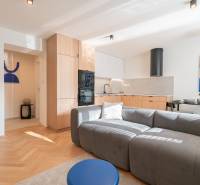 Living area of a 3-room apartment with a kitchen, gray sofa, and wood-patterned flooring.