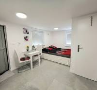 Interior of a cottage with a dining table, chairs, and a bed on a light floor.