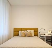 A bedroom in a studio apartment with a light-colored bed, curtains, and a white table with a lamp.