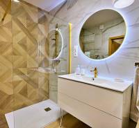 A bathroom in a 3-room apartment with a large illuminated mirror and gold accessories by the shower corner.