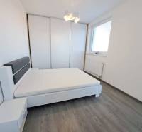 A bright bedroom in a two-room apartment with wood-patterned flooring, white furniture, and a built-in wardrobe.