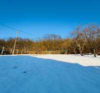 A snowy plot in Horna Ves surrounded by trees and a fence near a family house.