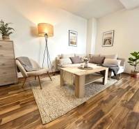 Living room in a three-room apartment with a wooden decor floor, light furniture, and a plant.