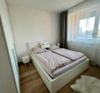 A bedroom with light furnishings, wood-patterned flooring, and a window in a 2-room apartment.