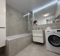 Bathroom in a 2-room apartment with a bathtub and a washing machine, light tiles and a sink.