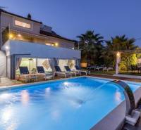 Villa in Kaštela with a lit pool, loungers, and palm trees in the garden at dusk.