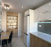 A kitchen in a 2-room apartment with white cabinets, a table, and wooden decor shelves.