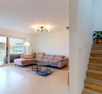 Interior of a family house: sofa, bookshelf, stairs with wood-patterned flooring, and a plant.