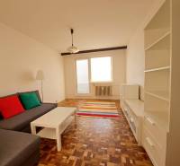 Living room in a two-room apartment with a sofa, white furniture, colorful pillows, and a wooden-patterned floor.