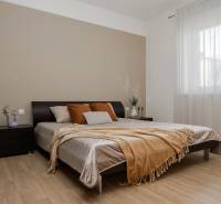 A bedroom in a 2-room apartment with a wood-patterned floor, a large bed, and a window with a sheer curtain.