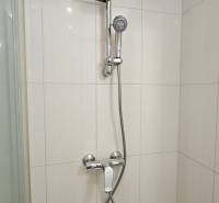 A shower corner in a studio apartment with white tiles and a chrome shower faucet.
