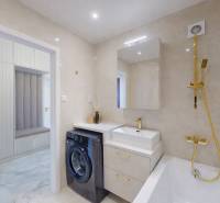 A bathroom in a 3-room apartment with a washing machine, sink, and bathtub with gold accessories.
