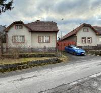A family house in Babiná with two neighboring houses, a parked car, and a stone fence.