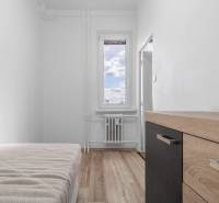 Jednoduchá izba s posteľou, komodou a podlahou s dreveným dekorom v 1-izbovom byte.