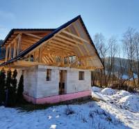An unfinished cabin on the street in Mýto pod Ďumbierom, surrounded by a snowy landscape and trees.