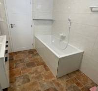 The bathroom in the family house has a bathtub, white tiles, and a floor with a wooden decor.