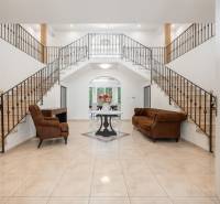 A spacious hall of a family house with a dominant staircase, brown furniture, and flowers.