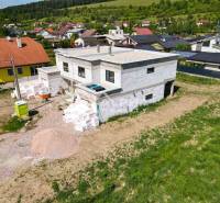 A family house under construction in Harichovce surrounded by other houses and green hills.