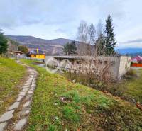 A building in Helcmanovce, surrounded by hills and a road with a lawn and trees.