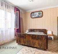 A room in a family house with a bed, a wooden-patterned floor, and patterned wallpaper.