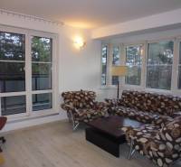 Living room in a two-room apartment with windows, wood-patterned flooring, and upholstered furniture.