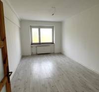 An empty room in a three-room apartment with a wood-patterned floor and a large window.