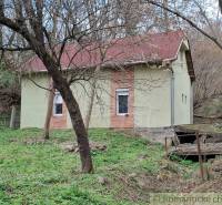 A cottage in Chľaba surrounded by deciduous trees and grass, with visible red roof shingles.