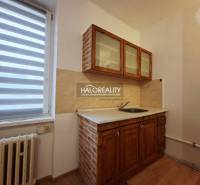 A kitchen in a studio apartment with wooden cabinets and blinds on the window.