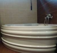A bathroom with a corner bathtub, shower, and shelving cabinet in a 2-room apartment.
