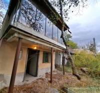 A cottage in Kamenín with a glass-enclosed balcony, surrounding nature, and an overgrown yard.