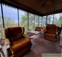 A veranda with chairs and a wooden floor, a cottage surrounded by greenery.