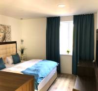 A studio apartment with light furniture, a bed, and blue curtains, with a wooden decor floor.