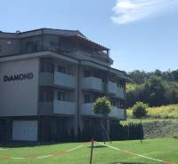 A 2-room apartment building in Podhájska surrounded by greenery and a cloudless sky.