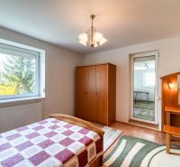 A bedroom in a three-room apartment with a wood-patterned floor, simple furniture, and a large window.