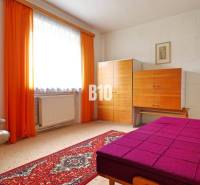 A room with a carpeted floor, a cream armchair, a colorful bed, and a wardrobe in a family house.