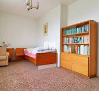 A room in a family house with a bed, a bookshelf, and an armchair on a carpet.