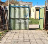 A yard by the cottage in Poltár with a gate and concrete paving under a blue sky.