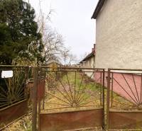 An old fence blocks the entrance to an overgrown plot. A family house with a white facade on the side.