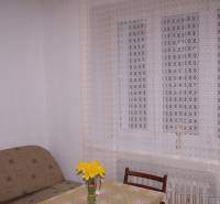 A room with a sofa, table, chair, and curtain, on the table a vase with flowers.