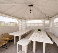 White gazebo with wooden furniture and rattan chairs in the garden.