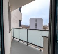 A balcony in a 2-room apartment with a view of the surrounding buildings through a glass railing.