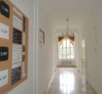 A hallway in offices with a decorative chandelier, white walls, and a board with company names.