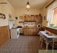 A kitchen in a family house with wooden furniture, tiled floor, and lace curtains.